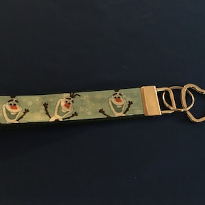 Könnte beinhalten: Ein blauer und grüner Schlüsselanhänger mit einem sich wiederholenden Muster von Olaf aus dem Film Frozen. Der Schlüsselanhänger hat einen goldenen Schlüsselring aus Metall mit einer herzförmigen Schlaufe.