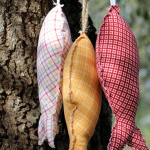 May include: Three fabric fish ornaments hanging from a tree branch. The fish are made of different patterned fabrics: red and white checkered, yellow and white striped, and white and red checkered.