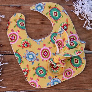 May include: A yellow baby bib with a colorful geometric pattern of turtles and circles. The bib has a matching fabric tie and a wooden teething ring.