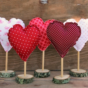 May include: Five fabric heart-shaped decorations on wooden sticks with wooden bases. The hearts are in various patterns of red, white, and pink.
