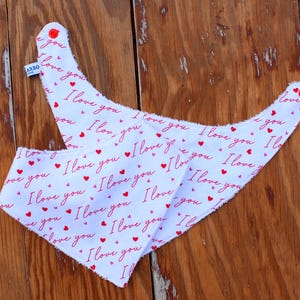 May include: Two white baby bibs with a repeating pattern of red hearts and the words "I love you" in red. The bibs have a triangular shape with rounded edges and red snap closures. The bibs are made of soft fabric.