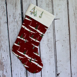 May include: A Christmas stocking with a burgundy background and a pattern of white lines, green trees, and snowflakes. The cuff is off-white with embroidered green Christmas trees. The stocking is made of fabric and is designed for the holiday season.