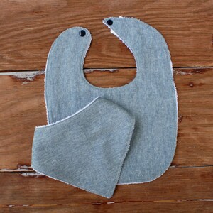 May include: Two-piece baby bib and burp cloth set. The bib is a light blue denim-like fabric with a rounded shape, snap closures, and white trim. The burp cloth is a matching triangular shape.