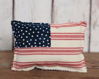 May include: A red, white and blue striped cushion with a blue star field in the top left corner. The cushion is made of fabric and has a slightly distressed look.