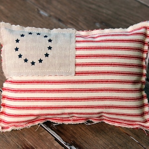 Op de afbeelding: Een rechthoekig, handgemaakt kussen met een patriottisch ontwerp. Het kussen heeft een crèmekleurige stof met rode strepen en een crèmekleurig vierkant met een cirkel van blauwe sterren, die doet denken aan de Amerikaanse vlag. De randen zijn gerafeld.