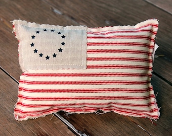 May include: A rectangular, handmade cushion with a patriotic design. The cushion features a cream-coloured fabric with red stripes and a cream-coloured square with a circle of blue stars, reminiscent of the American flag. The edges are frayed.