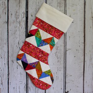 May include: A colorful patchwork Christmas stocking with a white cuff. The stocking is made of various fabrics in shades of pink, red, orange, yellow, green, blue, and black. The stocking is decorated with a geometric pattern of triangles.