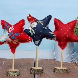 May include: Three fabric stars on wooden sticks, each with a different pattern. The stars are red, blue, and white with floral and geometric designs. The sticks are on wooden bases.