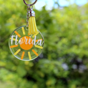 May include: A clear round keychain with a yellow tassel. The keychain features a yellow sun with orange rays and the word "Florida" in white lettering.