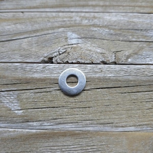 May include: A silver metal washer with a round hole in the center. The washer is sitting on a wooden surface.