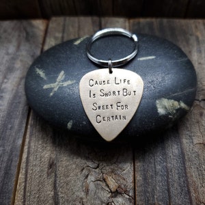 May include: A gold-colored guitar pick keychain with the text "Cause Life Is Short But Sweet For Certain".
