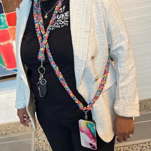 Puede incluir: Un cordón trenzado de colores con un llavero y una funda de teléfono adjuntos. El cordón lo lleva una persona que lleva una chaqueta blanca y pantalones negros. La funda del teléfono tiene un diseño abstracto de colores.
