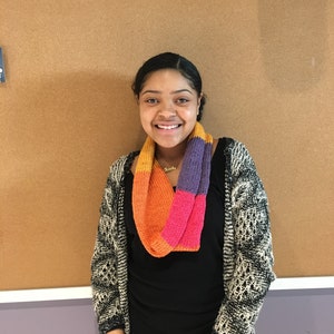 May include: A colorful knitted infinity scarf in shades of orange, pink, and purple. The scarf is worn around the neck and is paired with a black top and a patterned cardigan. The background is a corkboard.