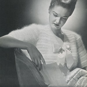 May include: A woman wearing a white fluffy knit cardigan with a satin bow. She is sitting in a chair and holding a piece of paper.