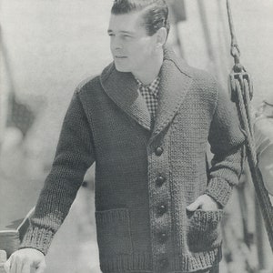 Op de afbeelding: Een grijze gebreide cardigan met een shawlkraag, een knoopsluiting en twee klepzakken. De cardigan is gemaakt van een dik, getextureerd garen.