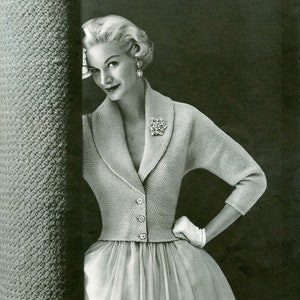 Może przedstawiać: Kobieta w jasnoszarym dzianinowym płaszczu z kołnierzem i guzikami. Płaszcz jest dopasowany i ma lekko rozkloszowaną spódnicę. Ma na sobie również białą spódnicę i białe rękawiczki. Obraz jest czarno-biały i zawiera tekst "Late Day Abbreviated Coat" u góry.