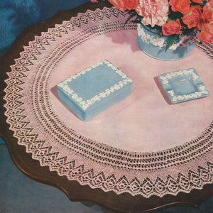 Op de afbeelding: Een vintage stilleven met een ronde, donkere houten tafel, bedekt met een roze tafelkleed en een delicate kanten rand. Op de tafel staan een lichtblauwe doos en asbak, beide met witte bloemmotieven. Een vaas met rozen en anjers staat op de achtergrond.