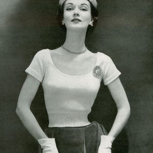 Op de afbeelding: Een zwart-witfoto van een vrouw in een witte top met korte mouwen, rok, handschoenen en een hoed. De top heeft een ronde hals en een decoratieve broche. De hoed is versierd met een kleine veer. De vrouw draagt een ketting en oorbellen.