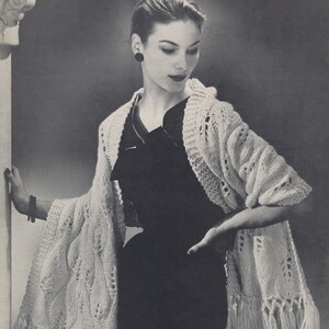 May include: A white crocheted shawl with fringe, worn over a black dress. The shawl has a lacy pattern and is draped over the shoulders of a woman.
