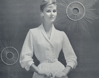 May include: A woman wearing a white crocheted dress with a double-breasted button closure and a belt at the waist. She is standing in front of a gray background with a decorative wall hanging behind her.