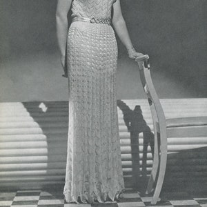 Könnte beinhalten: Eine Frau in einem weißen Häkelkleid mit Gürtel. Das Kleid hat einen gewellten Ausschnitt und kurze Ärmel. Sie steht neben einem Stuhl.