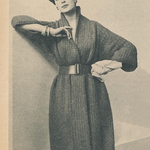 May include: A vintage black and white photograph of a woman wearing a long, textured coat with a wide belt. The coat has a shawl collar and is cinched at the waist with a large buckle. She wears a matching hat and bracelet.