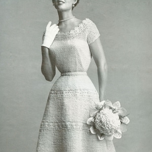 May include: A white crocheted wedding dress with a fitted bodice and a full skirt. The dress has a delicate lace pattern and is accented with a flower crown.