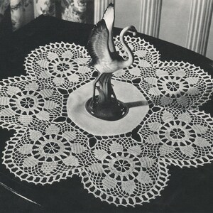 May include: A white crocheted doily with a delicate floral pattern. The doily is laid on a dark surface with a porcelain bird figurine in the center.