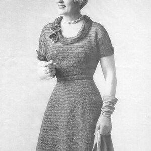 May include: A black and white photo of a woman wearing a knitted dress with a scoop neck and short sleeves. The dress has a textured pattern and a flower brooch on the neckline. The woman is wearing gloves and has her hair styled in a short bob.