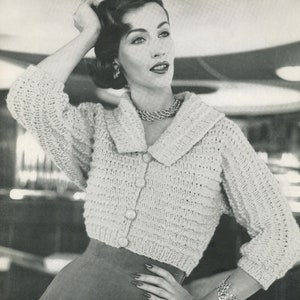 May include: A light beige, cropped knitted cardigan with a wide collar and three large buttons. The three-quarter sleeves and textured knit pattern add detail. The cardigan is paired with a skirt and jewelry.