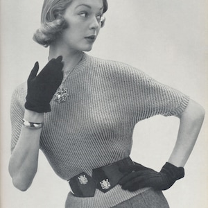May include: A woman wearing a gray knitted sweater with a black belt and black gloves. She is looking to the right of the frame.