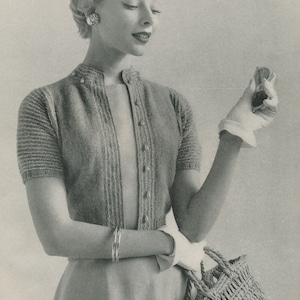 May include: A woman wearing a knitted cardigan with a button-up front and short sleeves. She is holding a woven straw handbag.