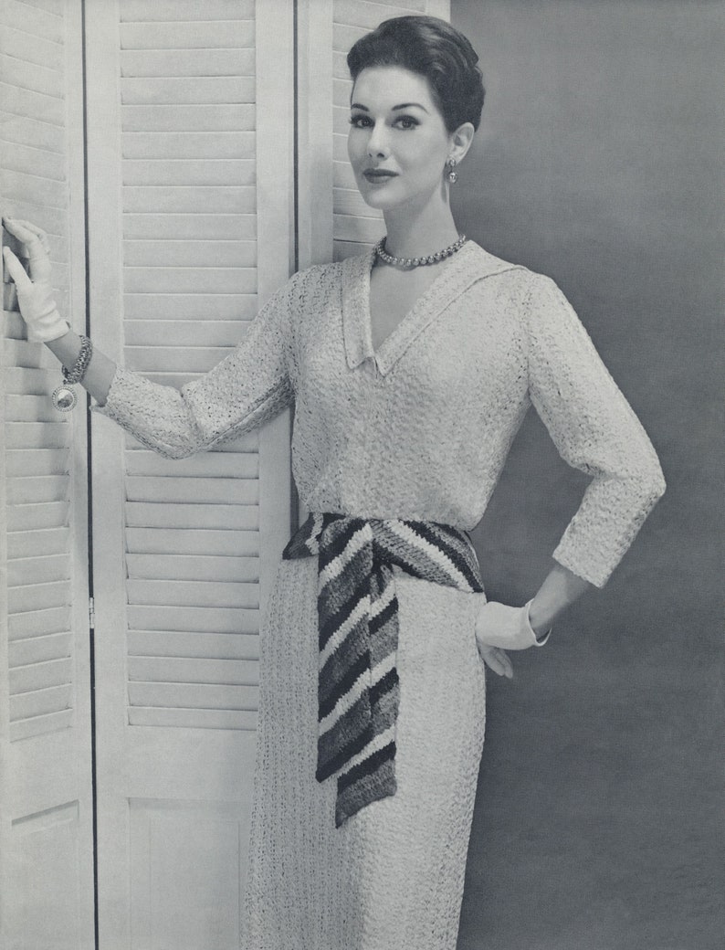 May include: A woman wearing a white crocheted dress with a striped crocheted belt. She is standing next to a white louvered door.