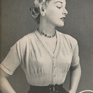 May include: A vintage-style, light-colored knit cardigan with short sleeves and a V-neckline. The cardigan is buttoned up and paired with a dark-colored skirt and a belt. The model is wearing a necklace and bracelet.
