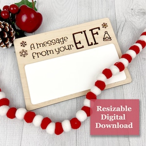 May include: A wooden sign with the text "A message from your ELF" and a blank white space for writing. The sign is decorated with snowflakes and a bell. A red and white pom-pom garland and Christmas decorations are in the background.