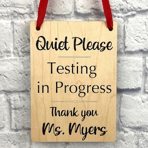Può includere: Un cartello di legno con il testo "Quiet Please Testing in Progress Thank you Ms. Myers" in lettere nere. Il cartello è appeso a un nastro rosso.
