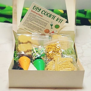 May include: A DIY cookie kit in a white box with instructions, cookie dough, sprinkles, and icing. The kit includes a tractor cookie cutter and green and gold sprinkles.