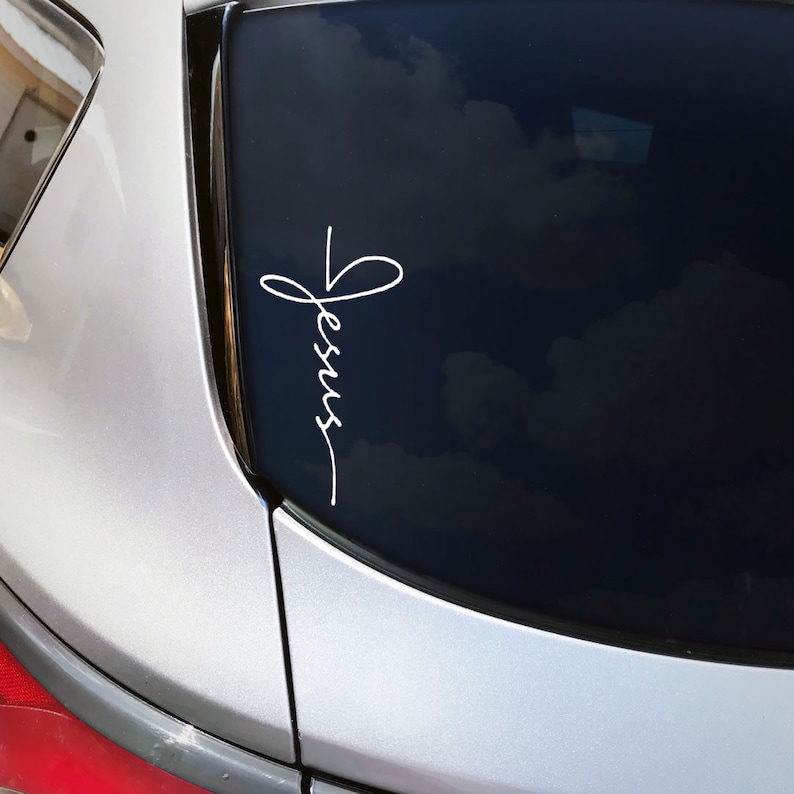 May include: A white vinyl decal with the word "Jesus" written in cursive script, with a cross formed by the "J" and the top of the "S". The decal is on the back window of a silver car.