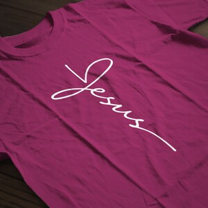 May include: A magenta t-shirt with the word "Jesus" written in white cursive script.