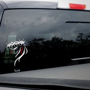 Puede incluir: Calcoman&iacute;a de vinilo blanco de una persona con una corona de espinas con sangre goteando por su rostro. La calcoman&iacute;a est&aacute; en la ventana trasera de una camioneta gris.