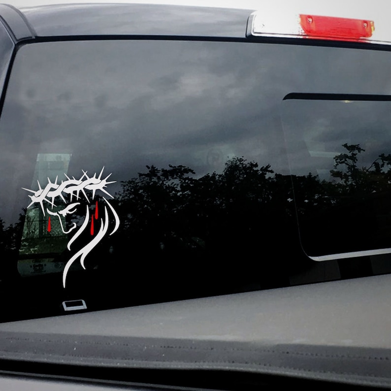 Puede incluir: Calcoman&iacute;a de vinilo blanco de una persona con una corona de espinas con sangre goteando por su rostro. La calcoman&iacute;a est&aacute; en la ventana trasera de una camioneta gris.
