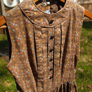 Puede incluir: Vestido vintage marrón con estampado de cuadrados multicolores. Cuello alto, pliegues y botones negros. Sin mangas, colgado de una percha de madera. La percha tiene la inscripción "New York City" y "7th Ave".