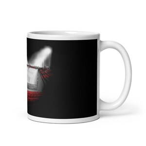 May include: White ceramic mug featuring a black and white illustration of a sports arena with red seating. The image is set against a black background, with a bright spotlight illuminating the arena.