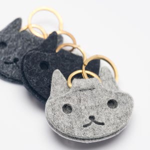 May include: Three felt cat-shaped keychains in shades of gray, each with a gold-colored keyring. The keychains have embroidered facial features, including eyes and a mouth. The keychains are arranged in a diagonal line.