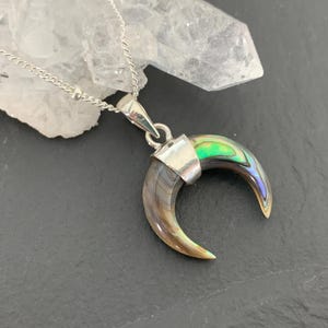 May include: A silver necklace featuring a crescent-shaped pendant. The pendant is made of iridescent abalone shell, displaying shades of green, blue, and brown. The necklace is displayed on a dark surface with a white crystal in the background.