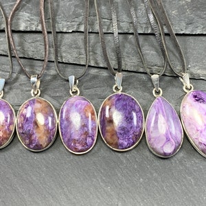 May include: Six silver pendants with purple and brown stone inserts on brown leather cords. The pendants are oval shaped and have a smooth, polished finish.