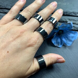 May include: Five black rings worn on a hand. The rings are all the same width and have a smooth, polished finish.