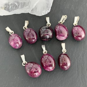 Peut inclure: Huit pendentifs en pierres précieuses polies, de forme ovale, rouge-violet foncé, avec des bélières argentées. Les pierres précieuses varient légèrement en taille et en forme, présentées sur une surface gris foncé.