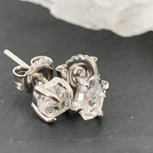 May include: A pair of silver stud earrings with clear, faceted gemstones. The earrings are set in a classic four-prong setting, and the posts are visible. The background is a dark surface with a white, blurred object.