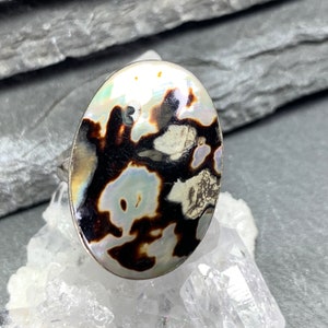 May include: Oval-shaped ring featuring a polished, iridescent stone with a black, brown, and white pattern. The ring has a silver band and is set against a neutral background.